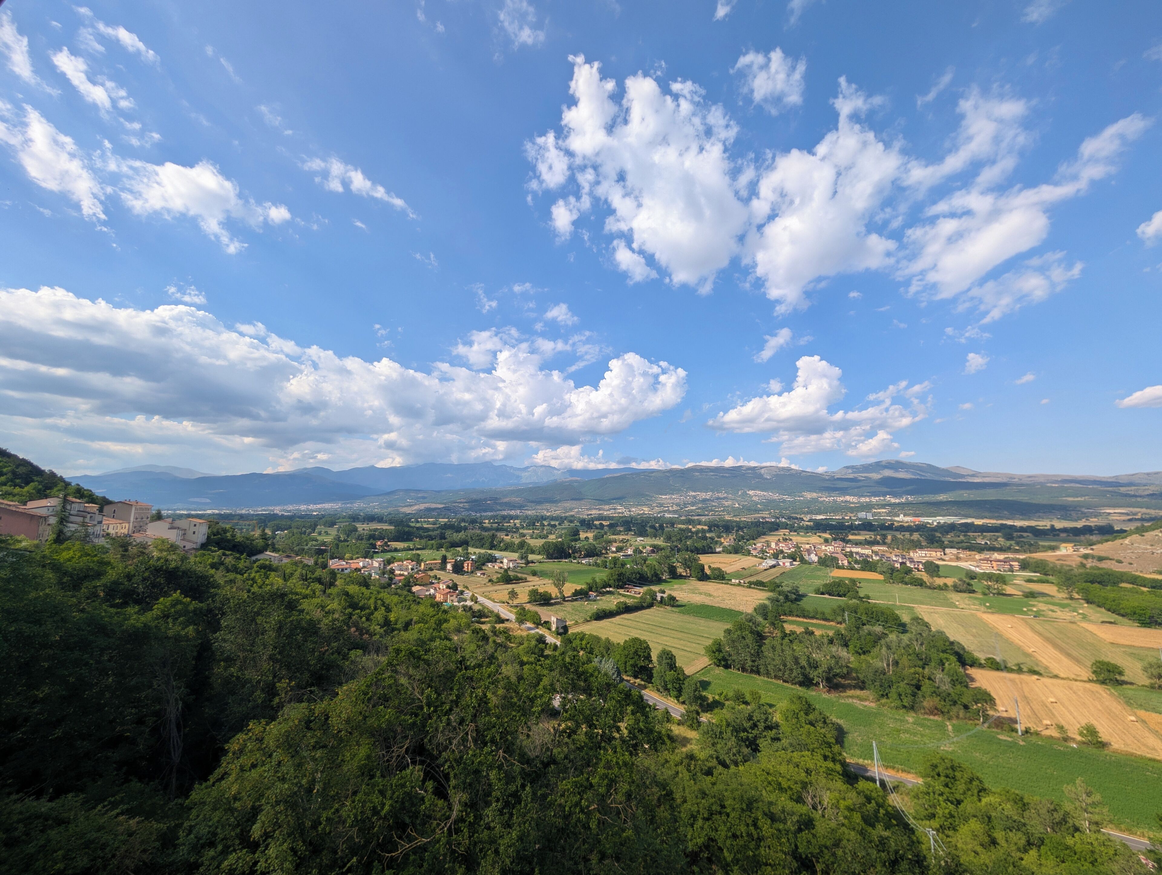 Ein weiter Blick über eine ländliche Landschaft mit einem Dorf, grünen Feldern und Bergen im Hintergrund.