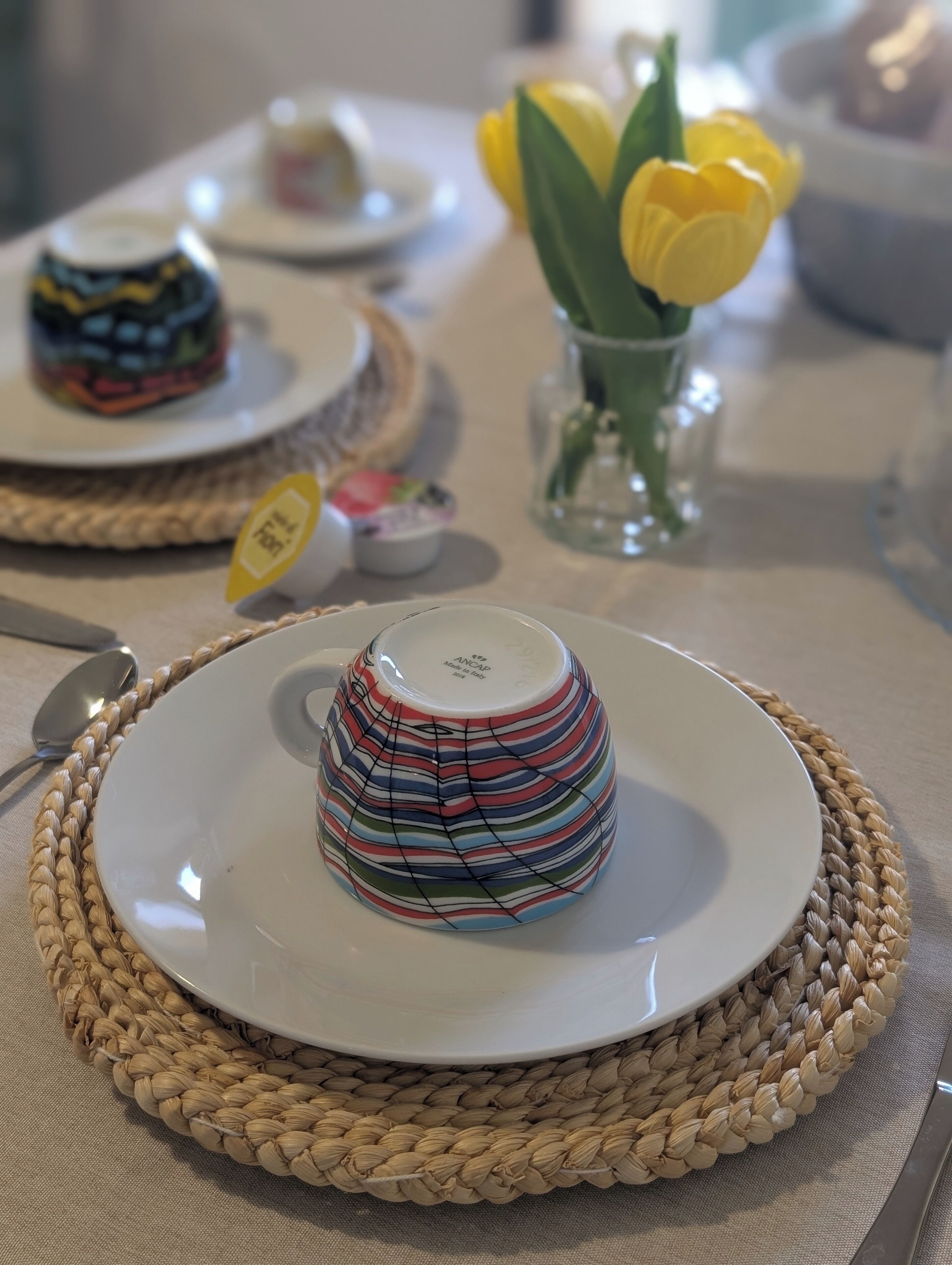 Una mesa con un posavasos tejido, un plato blanco y una taza de cerámica a rayas, además de un pequeño florero con flores amarillas.
