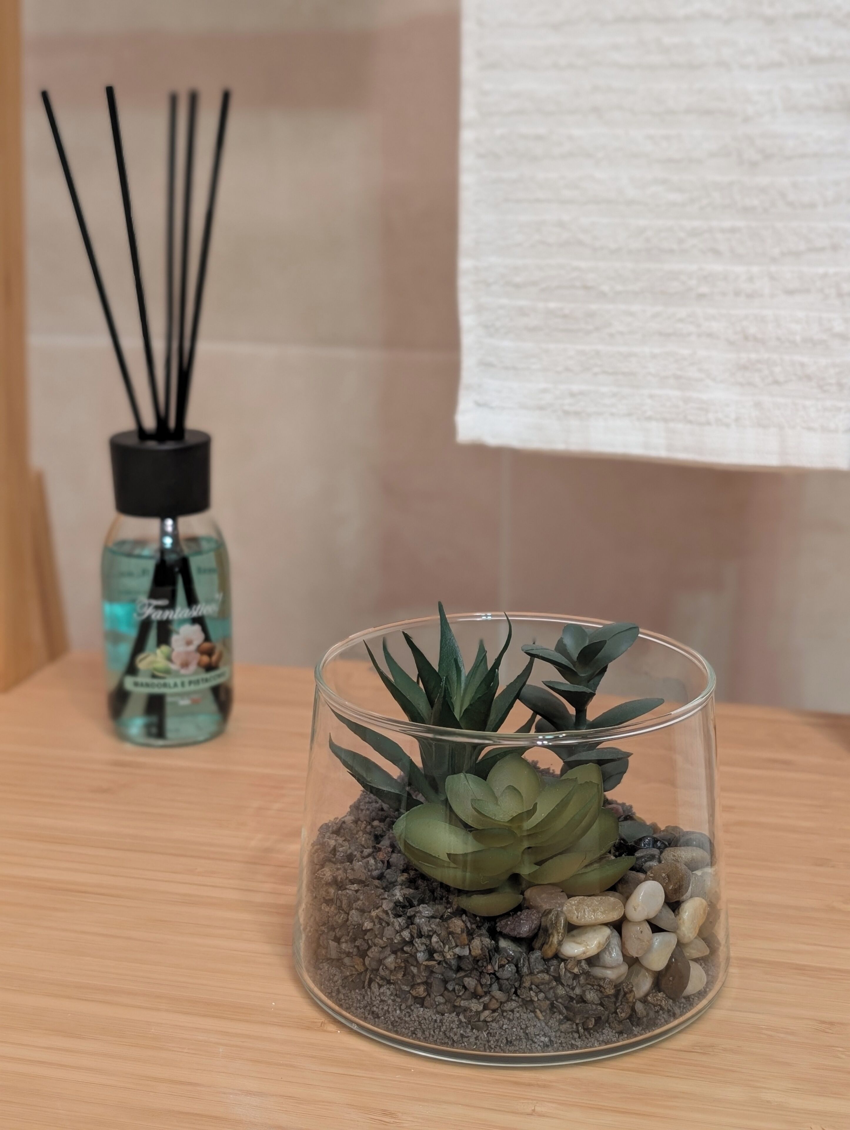 Un terrarium en verre contenant des plantes succulentes et des petits cailloux, posé sur une surface en bois, une bouteille avec des bâtonnets noirs, et une serviette blanche en arrière-plan.