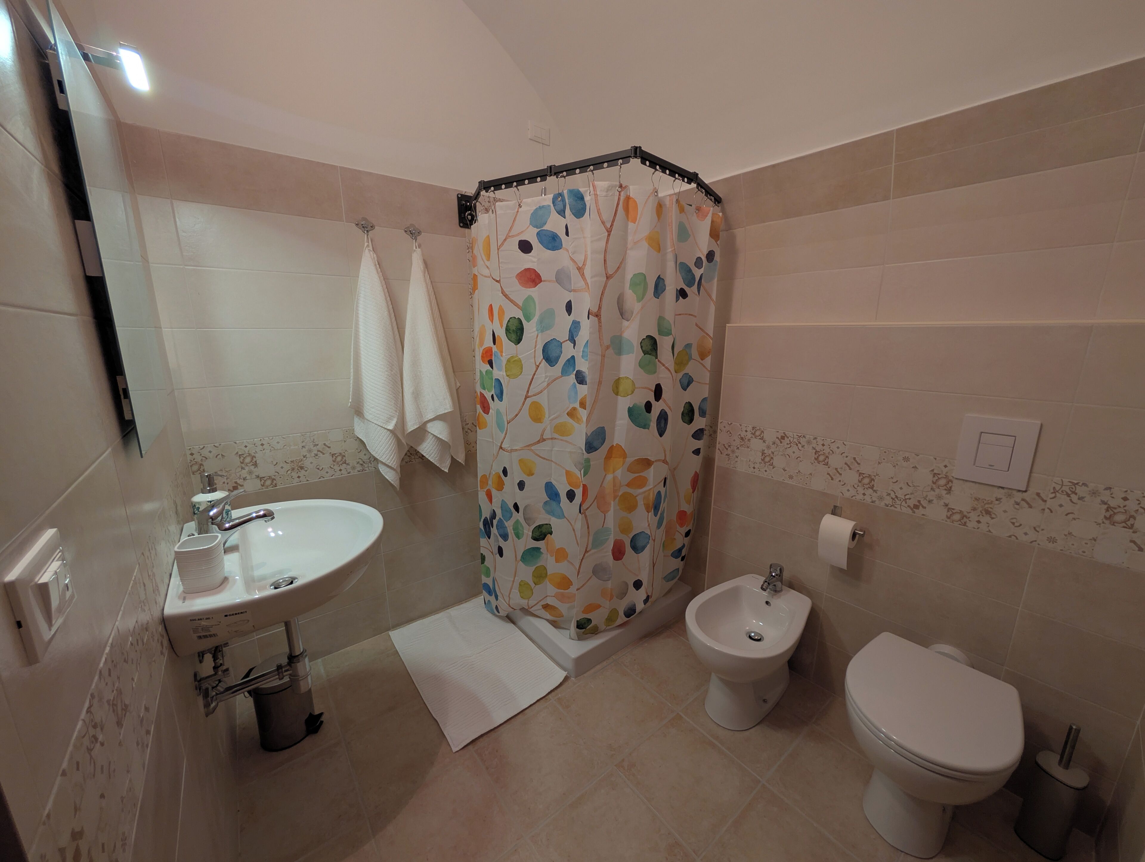 Een badkamer met een wastafel, bidet, toilet en een douchegordijn met een kleurrijk bladmotief.