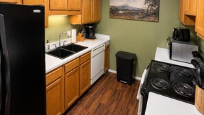 Une cuisine de taille réduite, avec des meubles de rangement en bois, un réfrigérateur noir, un évier, une cuisinière et une poubelle.