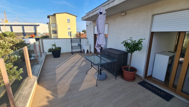 A rooftop terrace with a glass table, potted plants, a white cabinet, and a metal rack.