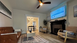Ein Wohnzimmer mit braunem Ledersofa, einem Holztisch, einem gemusterten Teppich, einem Kamin und einem an der Wand befestigten Fernseher.