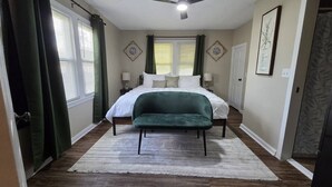 Quarto com uma cama, um banco verde, um ventilador de teto e uma janela com cortinas.