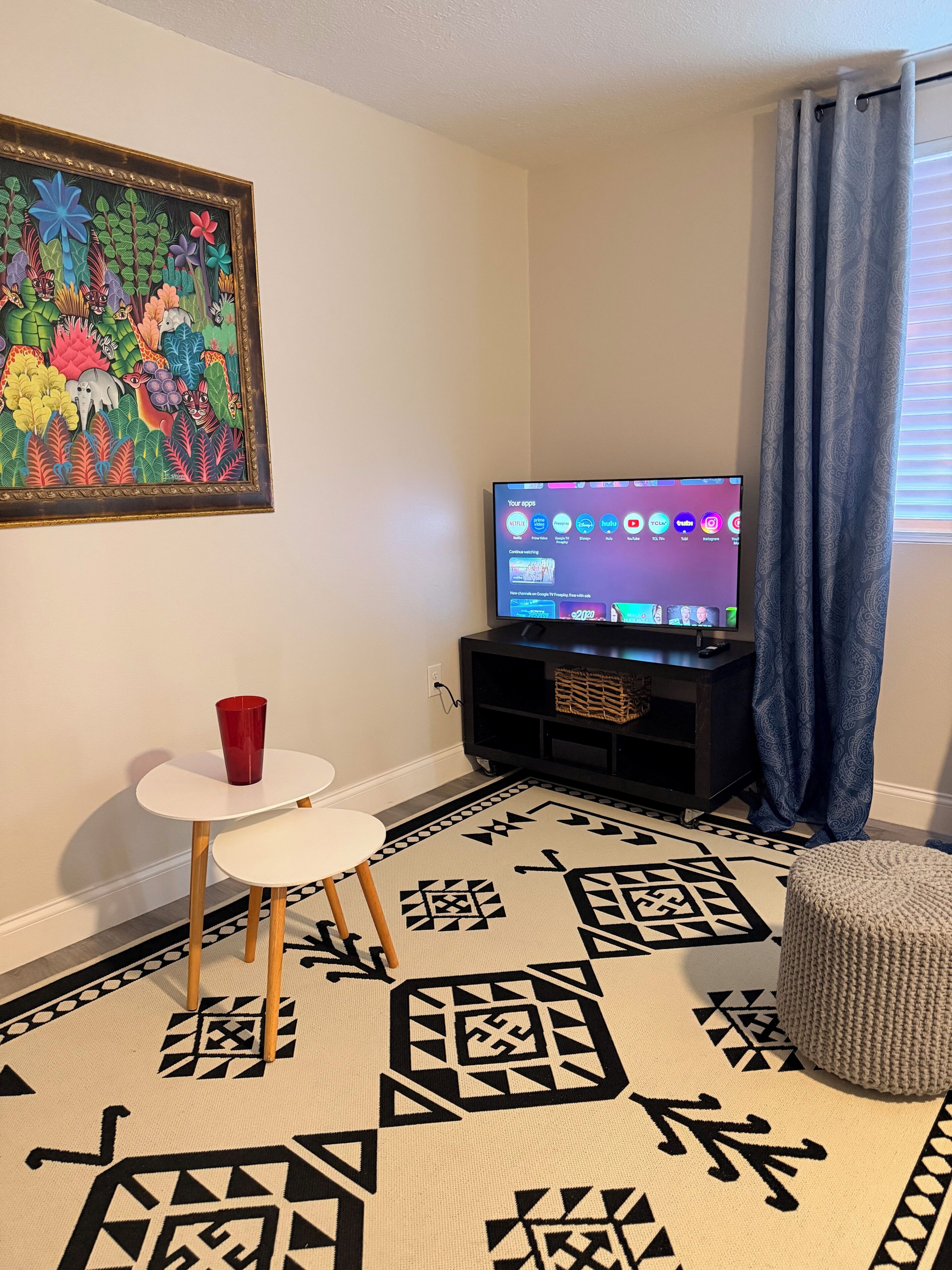 Una sala de estar con un televisor de pantalla plana, una mesita blanca con una taza roja, un tapete estampado y un cuadro colorido en la pared.
