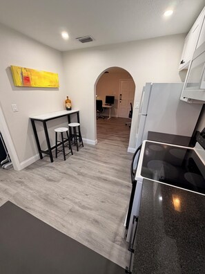 Una cocina moderna con encimera negra, un comedor con mesa blanca y dos taburetes, y un pasillo que conduce a una zona de estar.