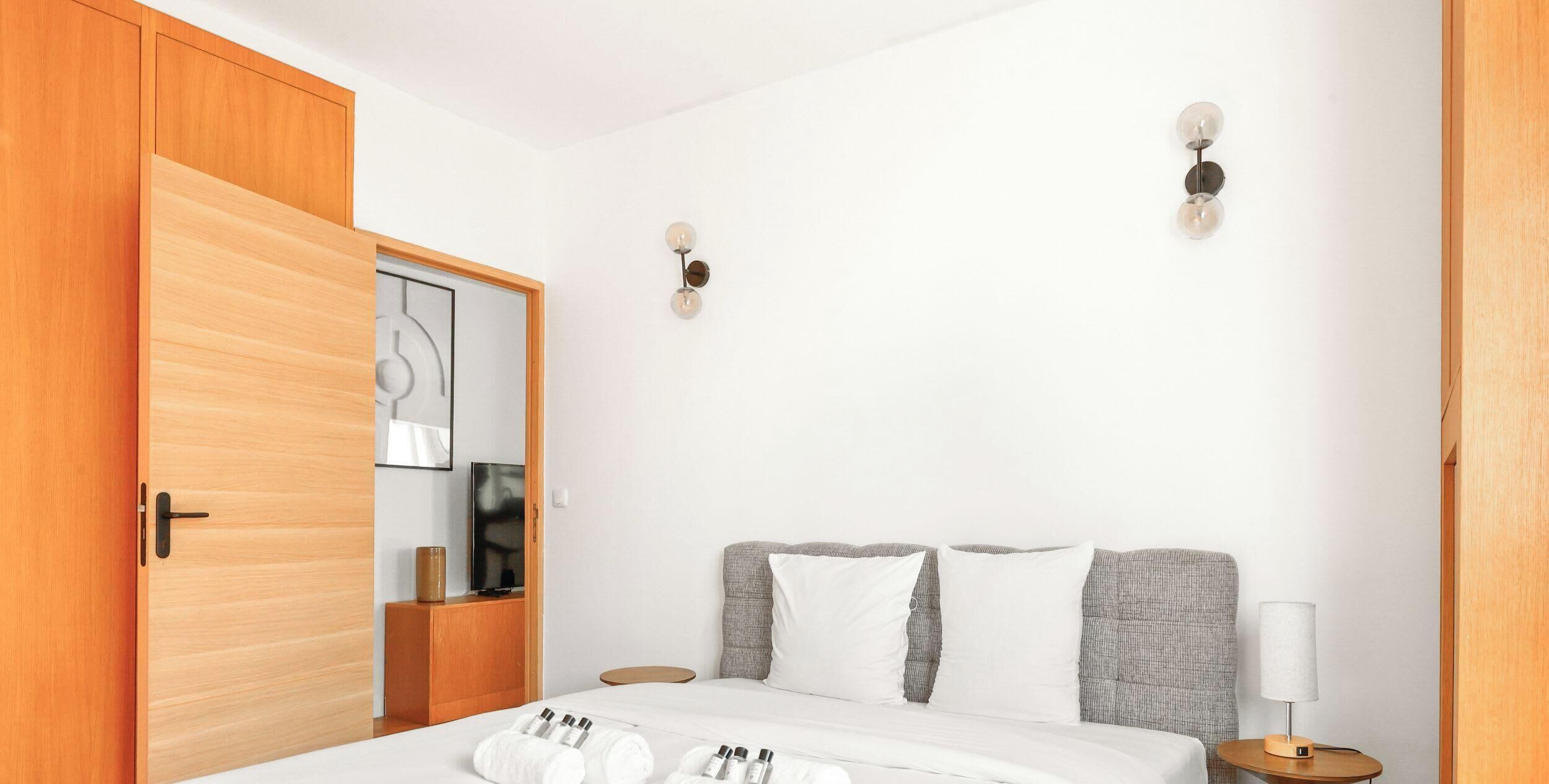 A hotel room with a bed, a wooden wardrobe, a mirror, a wall-mounted light fixture, and a bedside table with a lamp.