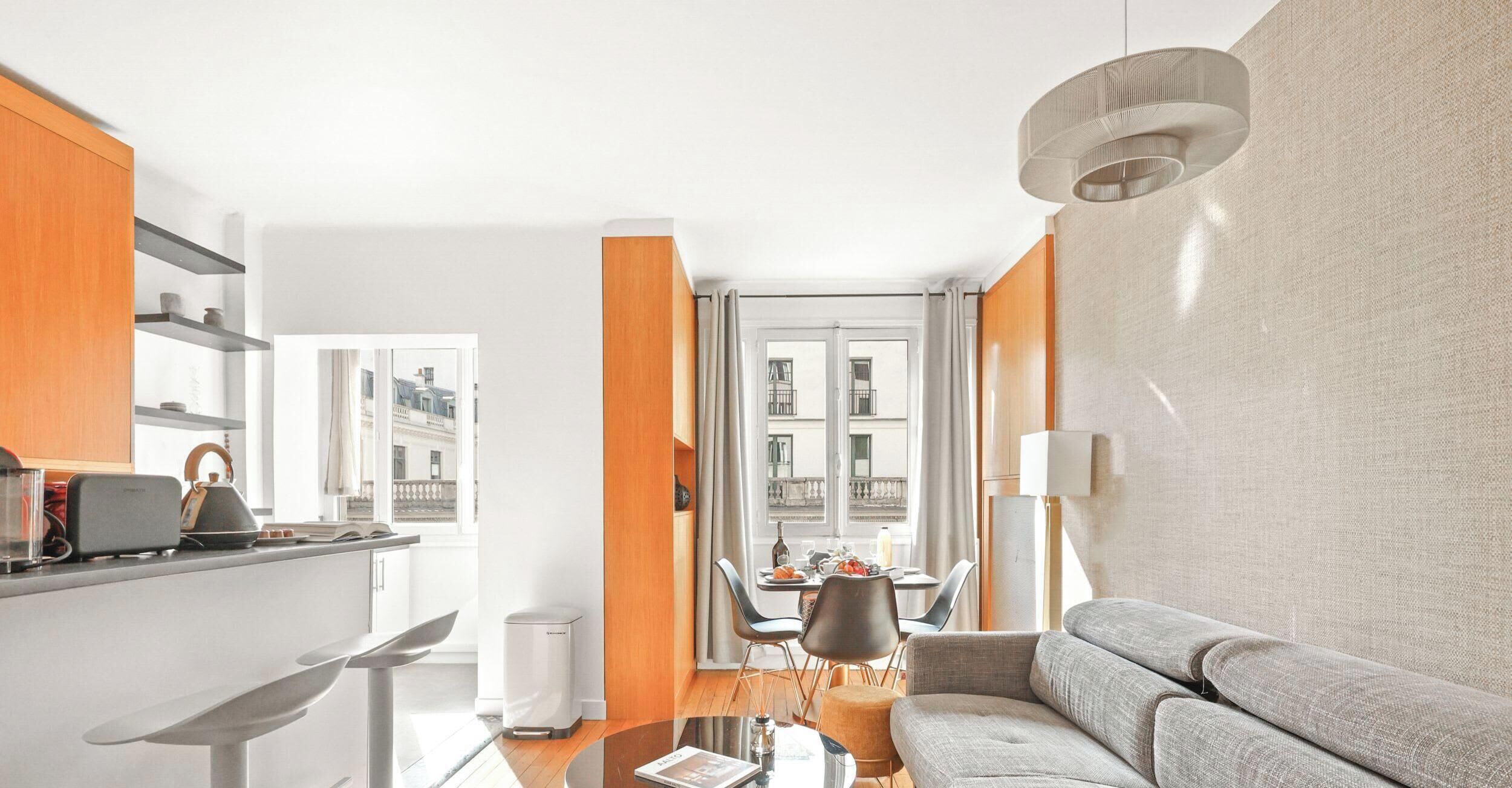 A modern living room with a sofa, a dining area with chairs, a kitchen with a counter, and a large window with a view of buildings.