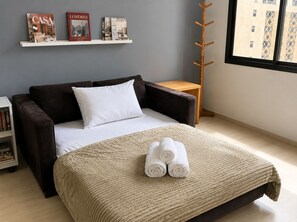 Quarto com cama, mesa de cabeceira, cabideiro de madeira e janela com vista para edifícios.