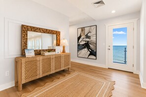 Un intérieur moderne avec une console en bois, un grand miroir, un tableau abstrait encadré et une porte donnant sur un balcon avec vue sur la mer.
