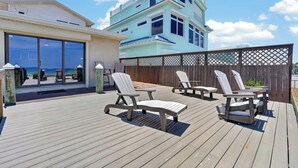A wooden deck with outdoor furniture, a view of the ocean, and a building with a light blue facade. - Beautiful 4-bedroom beach front house in Ponce Inlet (Port Orange)