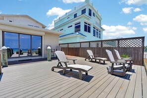Eine Holzterrasse mit Gartenmöbeln, Blick auf den Ozean und ein Gebäude mit hellblauer Fassade.