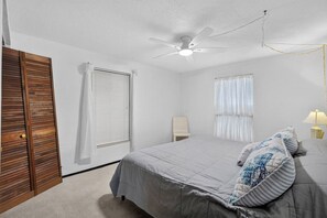 Ein Schlafzimmer mit einem Bett, einem Deckenventilator, einem Nachttisch mit Lampe, einem Sessel und einem Fenster mit Jalousien.