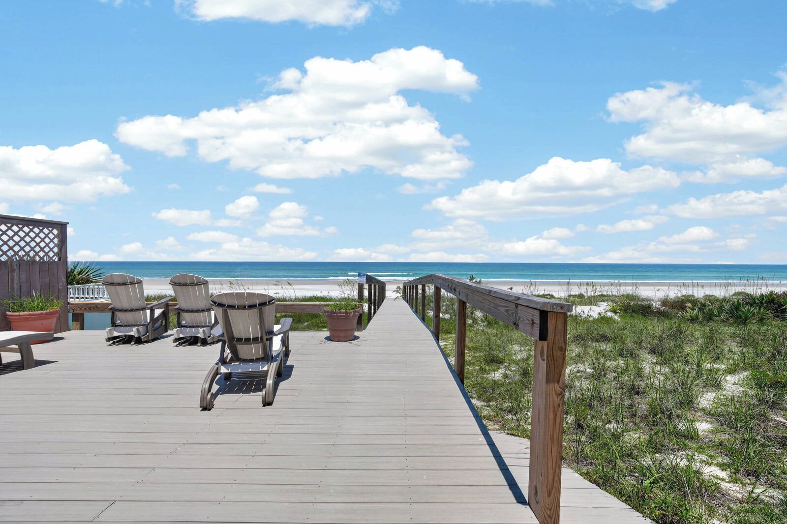 A wooden deck with chairs and potted plants, leading to a beach with a wooden railing.