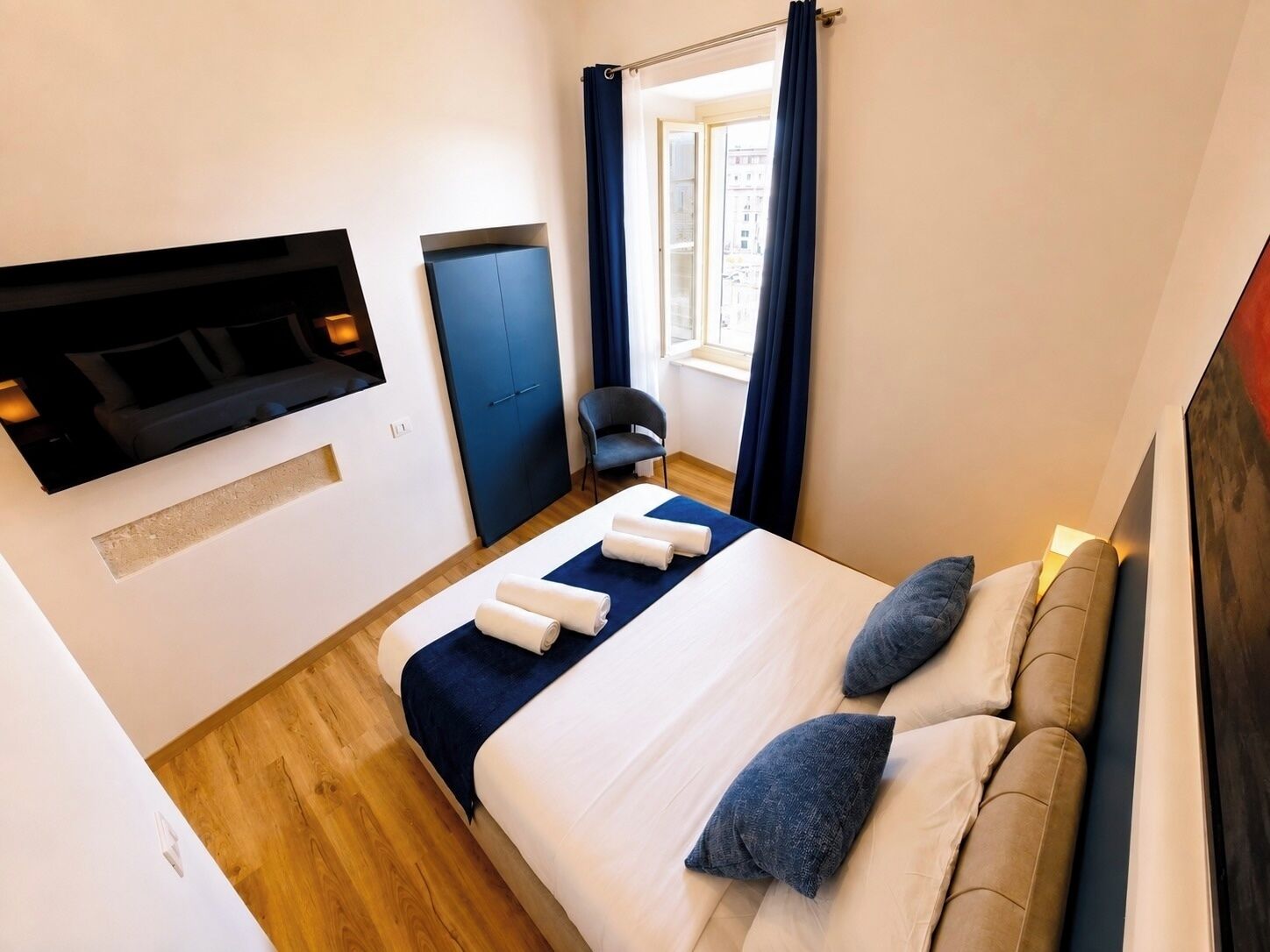 Ein Hotelzimmer mit einem Bett, einem an der Wand befestigten Fernseher, einem Fenster mit Vorhängen und einem Sessel.