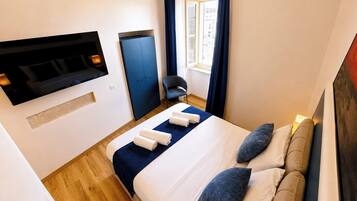 Ein Hotelzimmer mit einem Bett, einem an der Wand befestigten Fernseher, einem Fenster mit Vorhängen und einem Sessel.