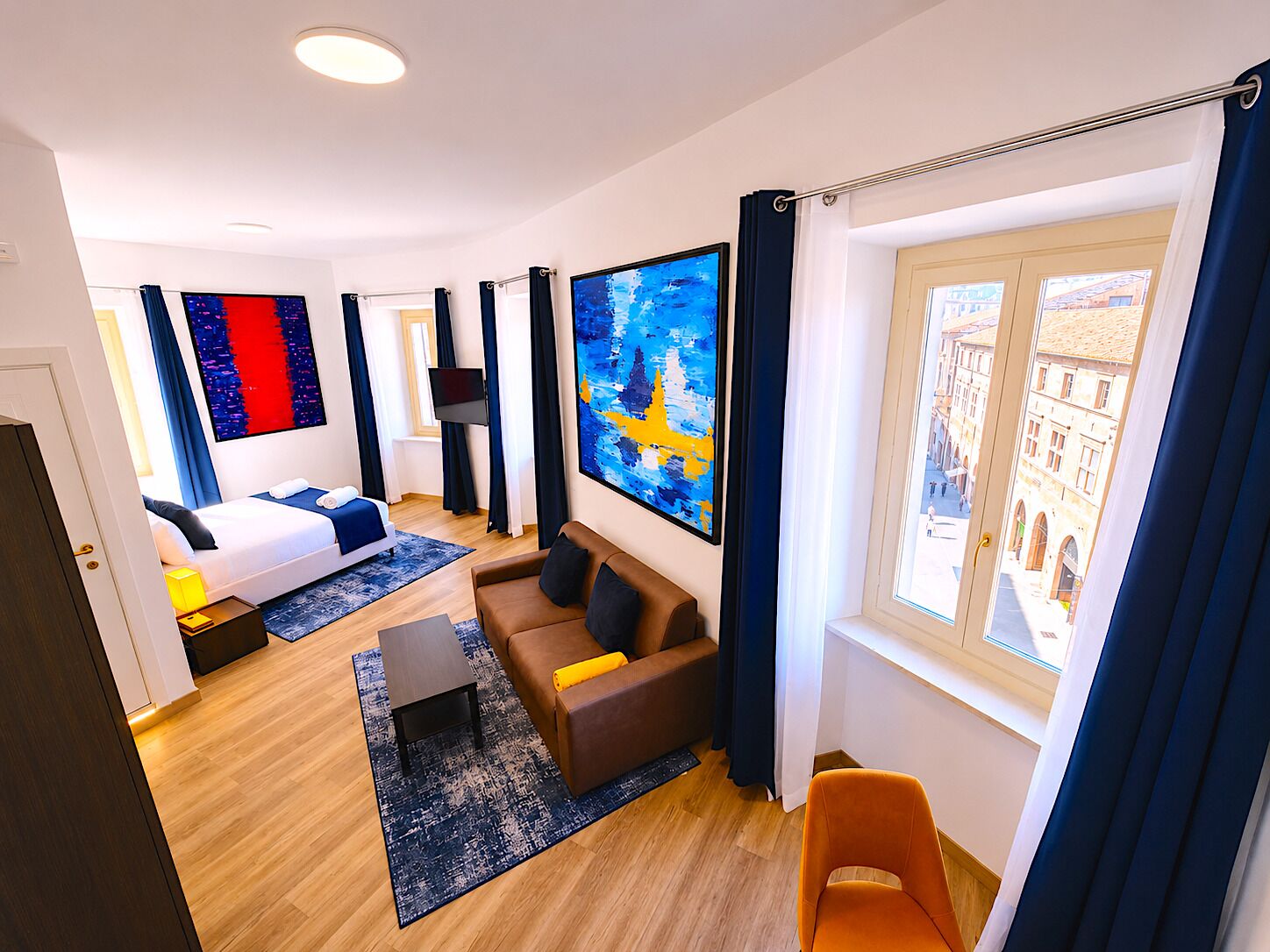Una habitación de hotel moderna con cama, sofá, televisor y un amplio ventanal con vista a la ciudad.