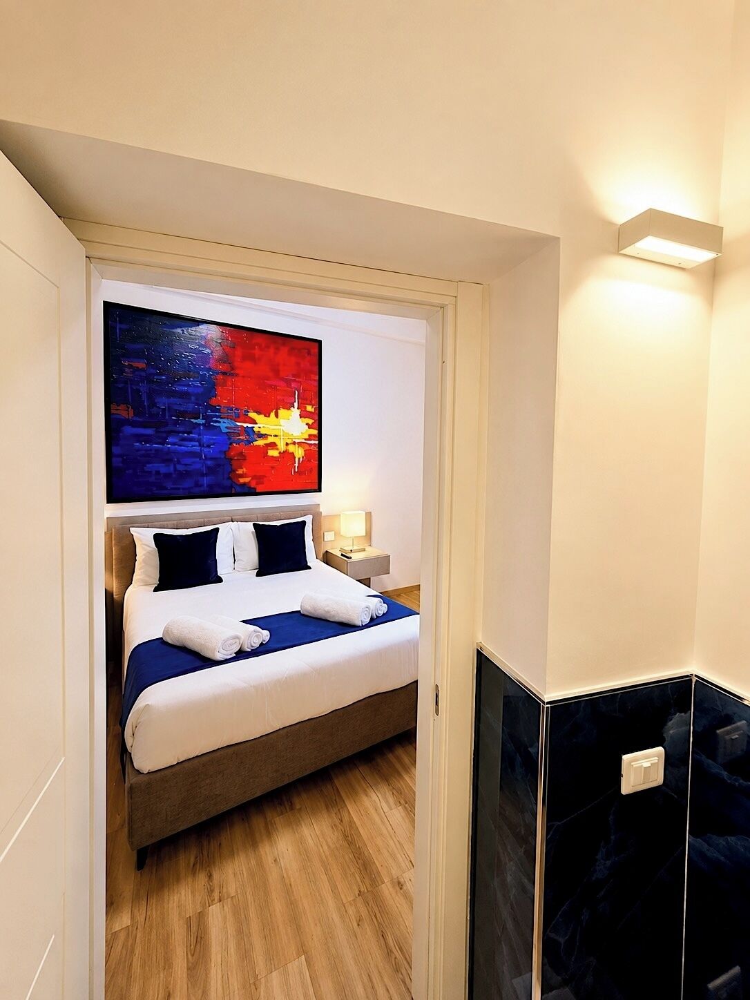 Une chambre d’hôtel avec un lit, une table de chevet, un tableau accroché au mur et un coin avec un meuble de couleur sombre.