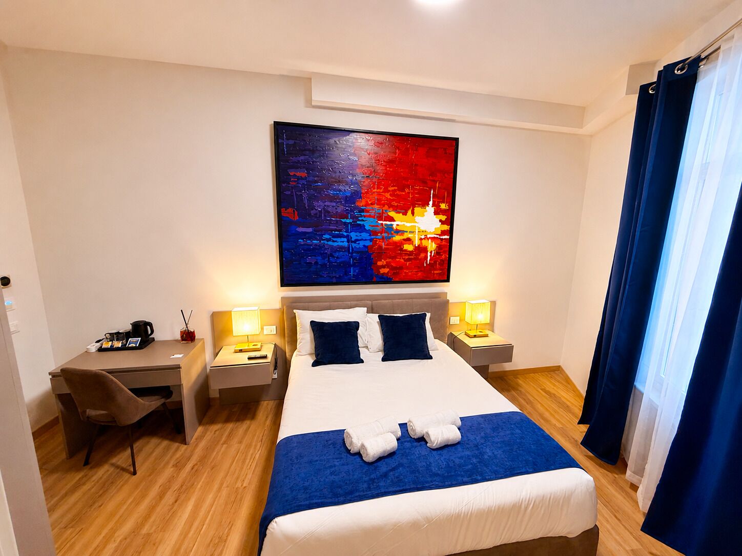 Una camera d'albergo con un letto grande, due comodini con lampade, una scrivania con una sedia e un grande quadro astratto appeso al muro.