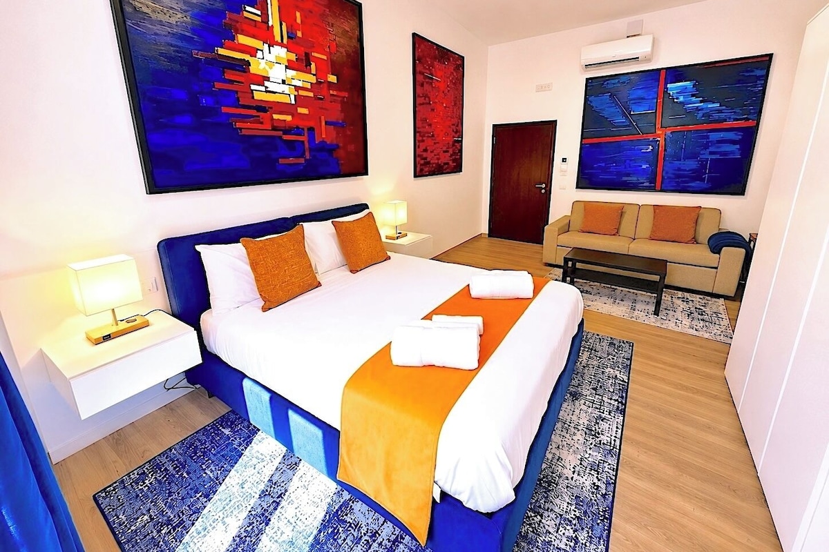 A hotel room with a large bed, a sofa, a desk, and two paintings on the walls.