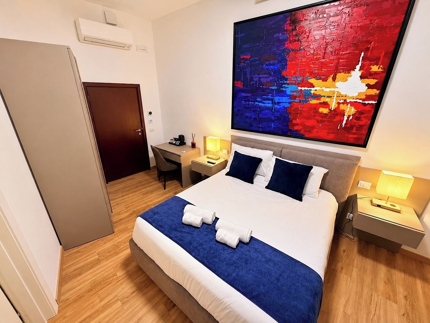 A hotel room with a bed, a desk, a chair, a nightstand, a lamp, and a large abstract painting on the wall.