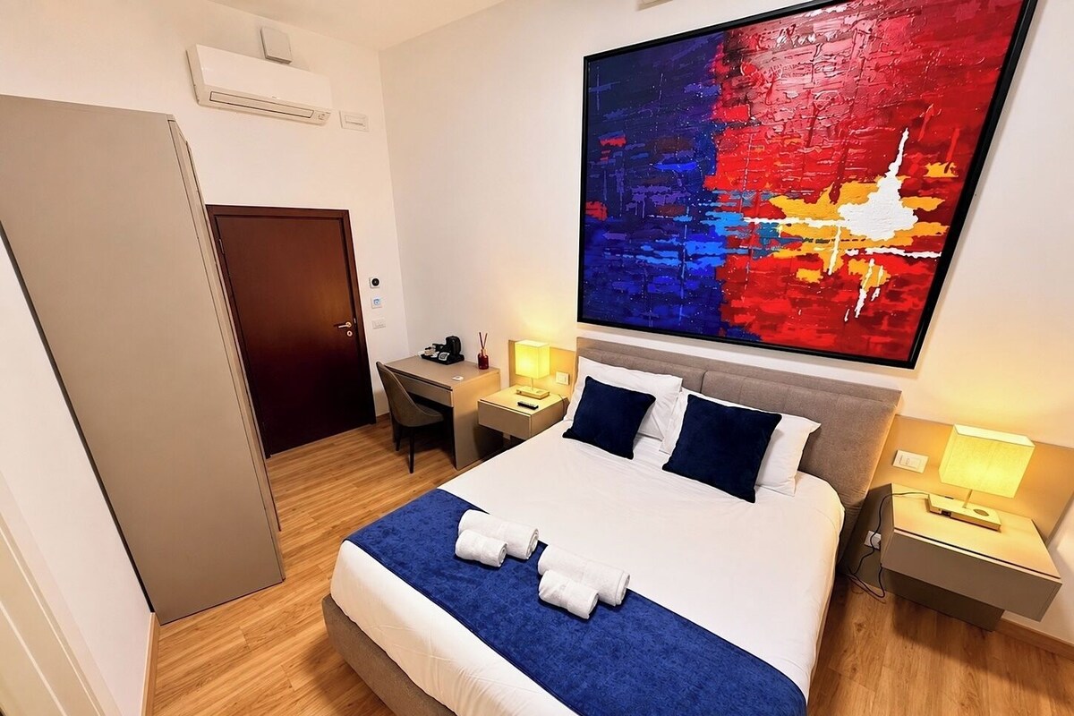 Habitación de hotel con cama, escritorio, silla, mesita de noche, lámpara y un cuadro abstracto grande en la pared.