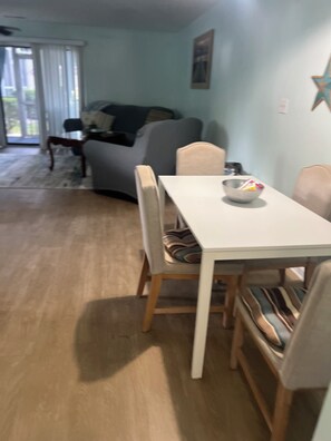 Ein Essbereich mit einem weißen Tisch, Stühlen und einer Schüssel auf dem Tisch. Im Hintergrund befinden sich eine Couch und ein Fenster.