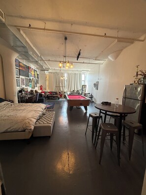 A room with a bed, a pool table, a dining table with chairs, and a refrigerator. - Brooklyn Loft with 8 ft pool table, king sized bed, full wall of windows and AC! (brooklyn)