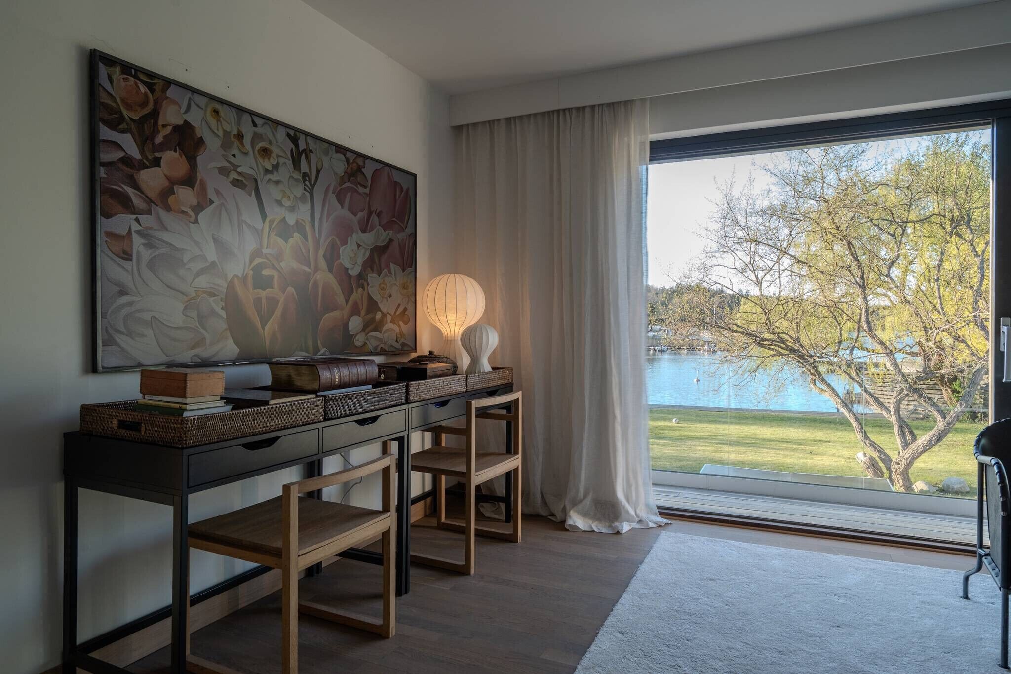 Ett rum med ett stort fönster, en tavla, en lampa och utsikt över träd och en vattenyta.