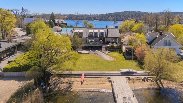 A modern house with a large deck overlooking a river, surrounded by trees and a clear view of a lake.
