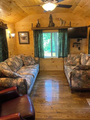 Interieur van een houten chalet met twee banken met bloemenprint, een leren fauteuil, een plafondventilator, een tv aan de muur en een raam met gordijnen.