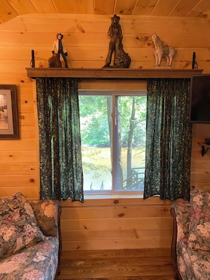 A cabin interior with a wooden ceiling, a window with curtains, a mounted statue, and floral-patterned furniture.