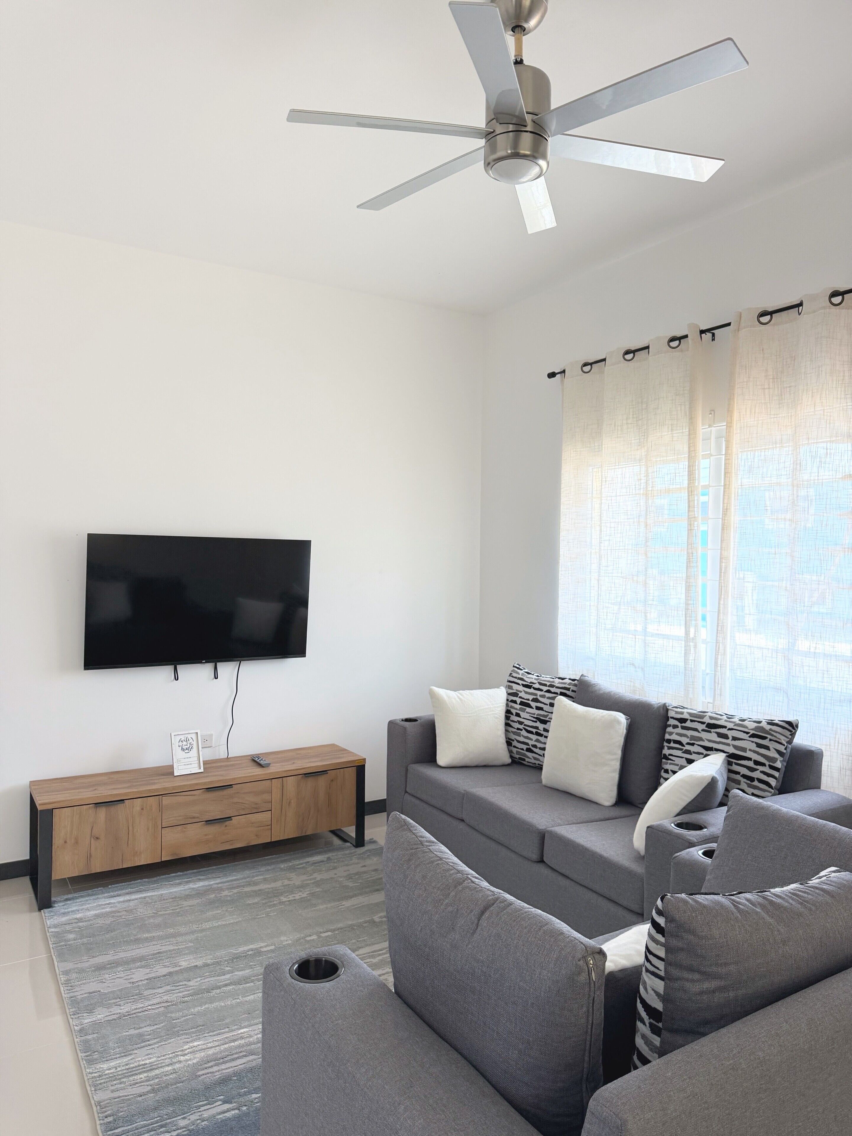 A modern living room with a flat-screen TV, grey sofa, wooden side table, and a ceiling fan.