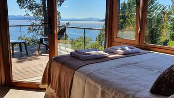 Un dormitorio con un ventanal que da a un lago y montañas, una cama con una manta y una silla en una terraza.