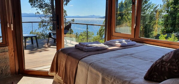 A bedroom with a large window overlooking a lake and mountains, a bed with a blanket, and a chair on a deck.