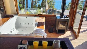 Bandeja de desayuno con tostadas, mantequilla, mermelada y jugo, jacuzzi y estufa de leña con vista al lago.