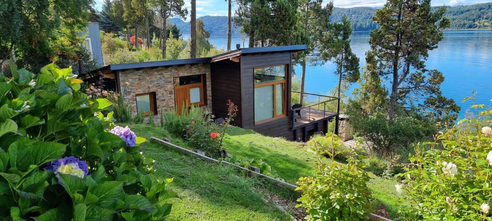 Una cabaña moderna con terraza con vistas a un lago, rodeada de vegetación exuberante.