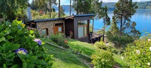A modern cabin with a deck overlooking a lake, surrounded by lush greenery.