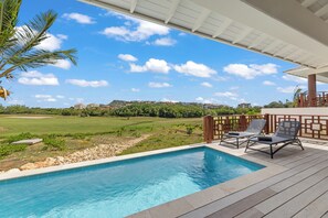 Ein Schwimmbad mit Liegestühlen, einer überdachten Terrasse und Blick auf einen Golfplatz und Gebäude.