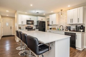 A modern kitchen with white cabinets, a central island with bar stools, stainless steel appliances, and a dishwasher. - Downtown Oasis! Hot Tub, 2 King Beds, Garage Parking, Huge Private Yard! (Milwaukee)