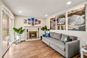 A living room with a fireplace, a grey sofa, and a large window. - Downtown Oasis! Hot Tub, 2 King Beds, Garage Parking, Huge Private Yard! (Milwaukee)