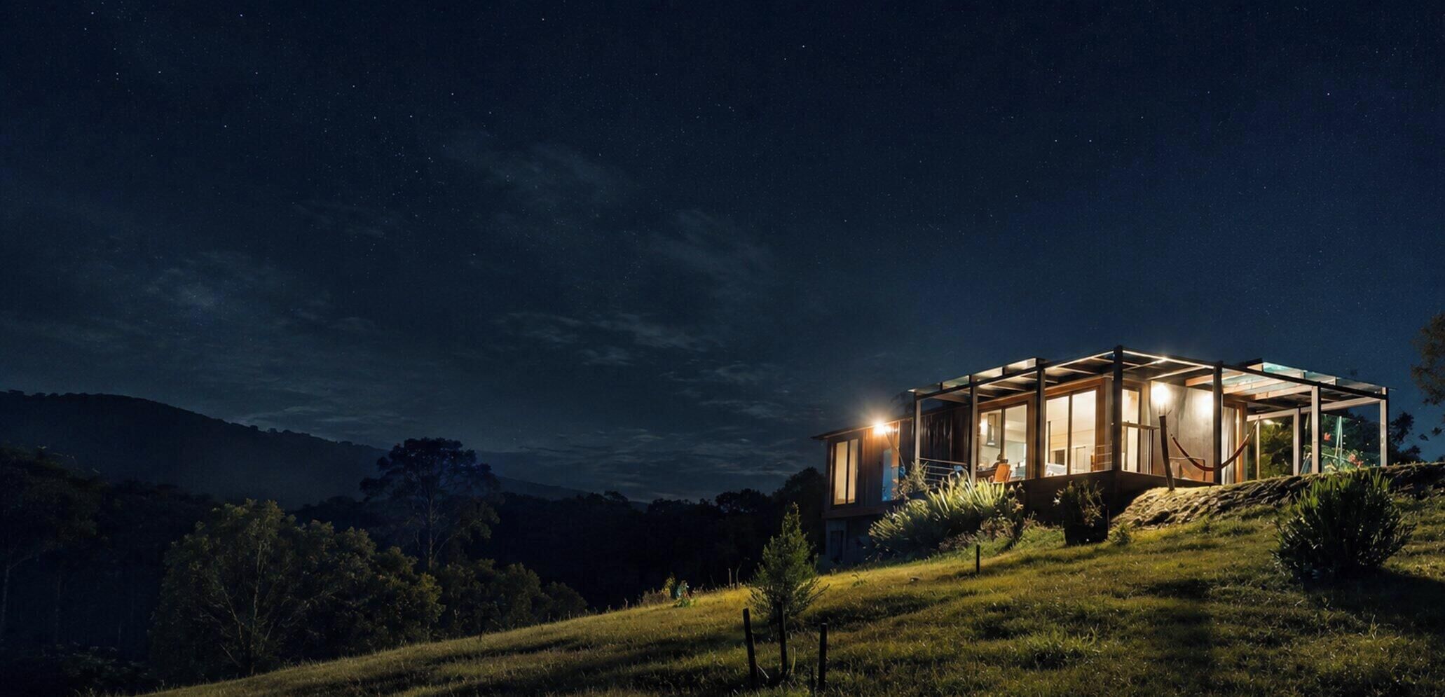 A modern house with a glass facade, situated on a grassy hill with a starry night sky.