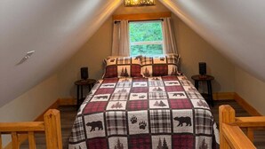 A bedroom with a sloped ceiling, a bed with a quilted blanket, a window with curtains, and a small table with a lamp. - Bear Hollow 2-bed cabin in charming Old Fort with AC (Old Fort)