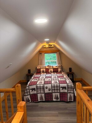 Ein Schlafzimmer mit schräg verlaufender Decke, einem Bett mit gesteppter Decke, einem Fenster mit Vorhängen und einem kleinen Tisch mit einer Lampe.