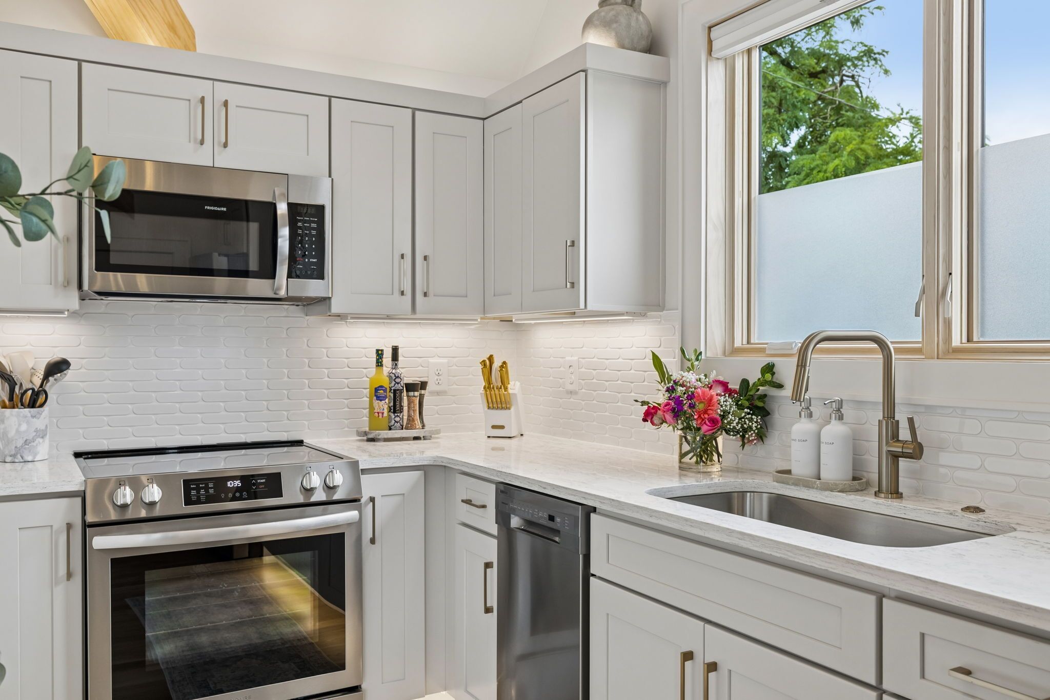 A modern kitchen with white cabinetry, stainless steel appliances, a brick backsplash, and a window providing natural light.