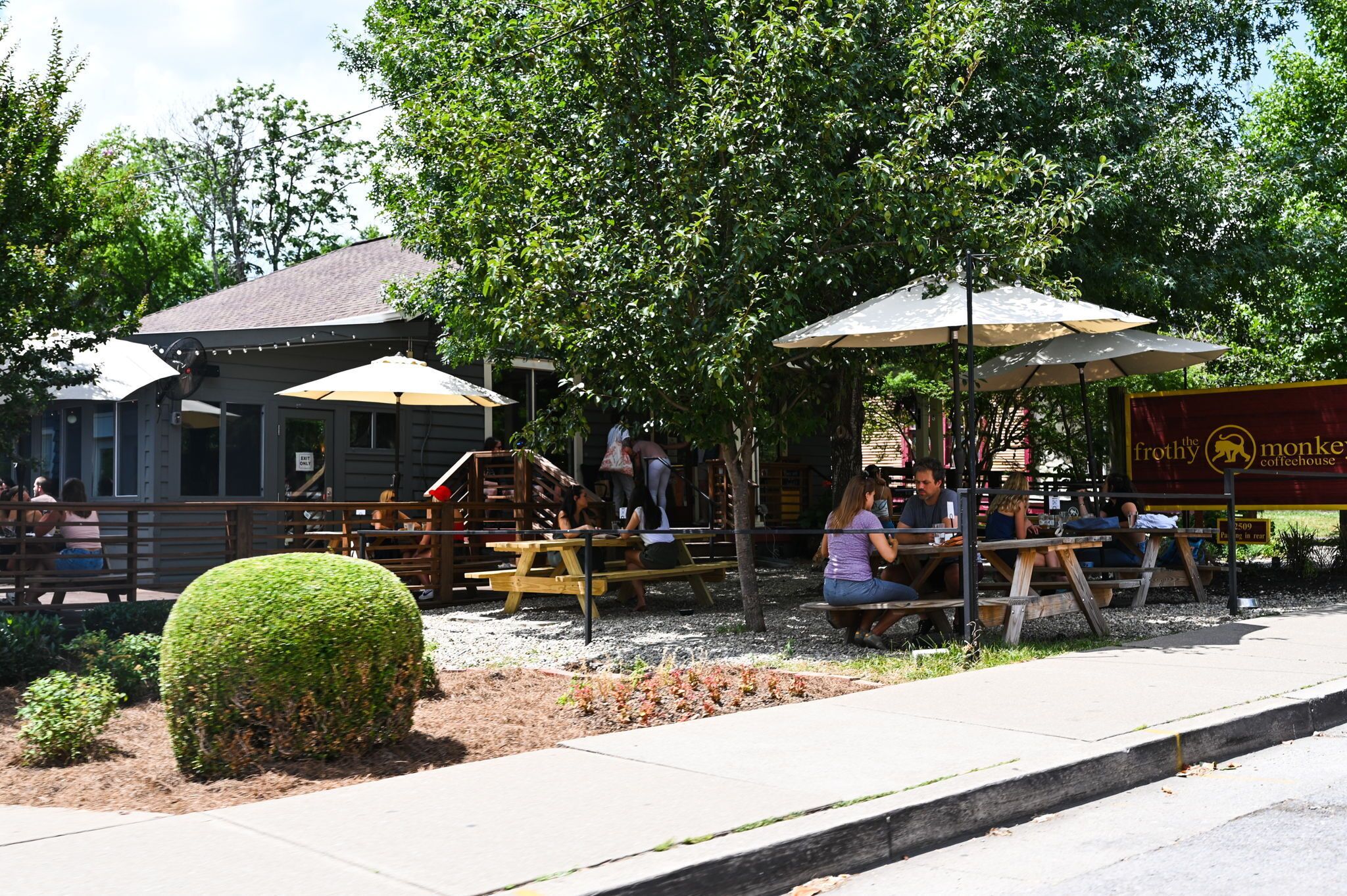 An outdoor seating area with tables and umbrellas, a building with a sign, and a well-maintained hedge.