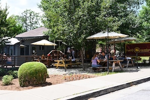 An outdoor seating area with tables and umbrellas, a building with a sign, and a well-maintained hedge.