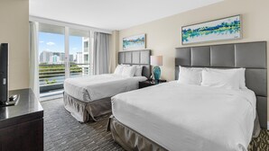 Quarto de hotel com duas camas, televisão, escrivaninha e vista para a cidade através de amplas janelas.