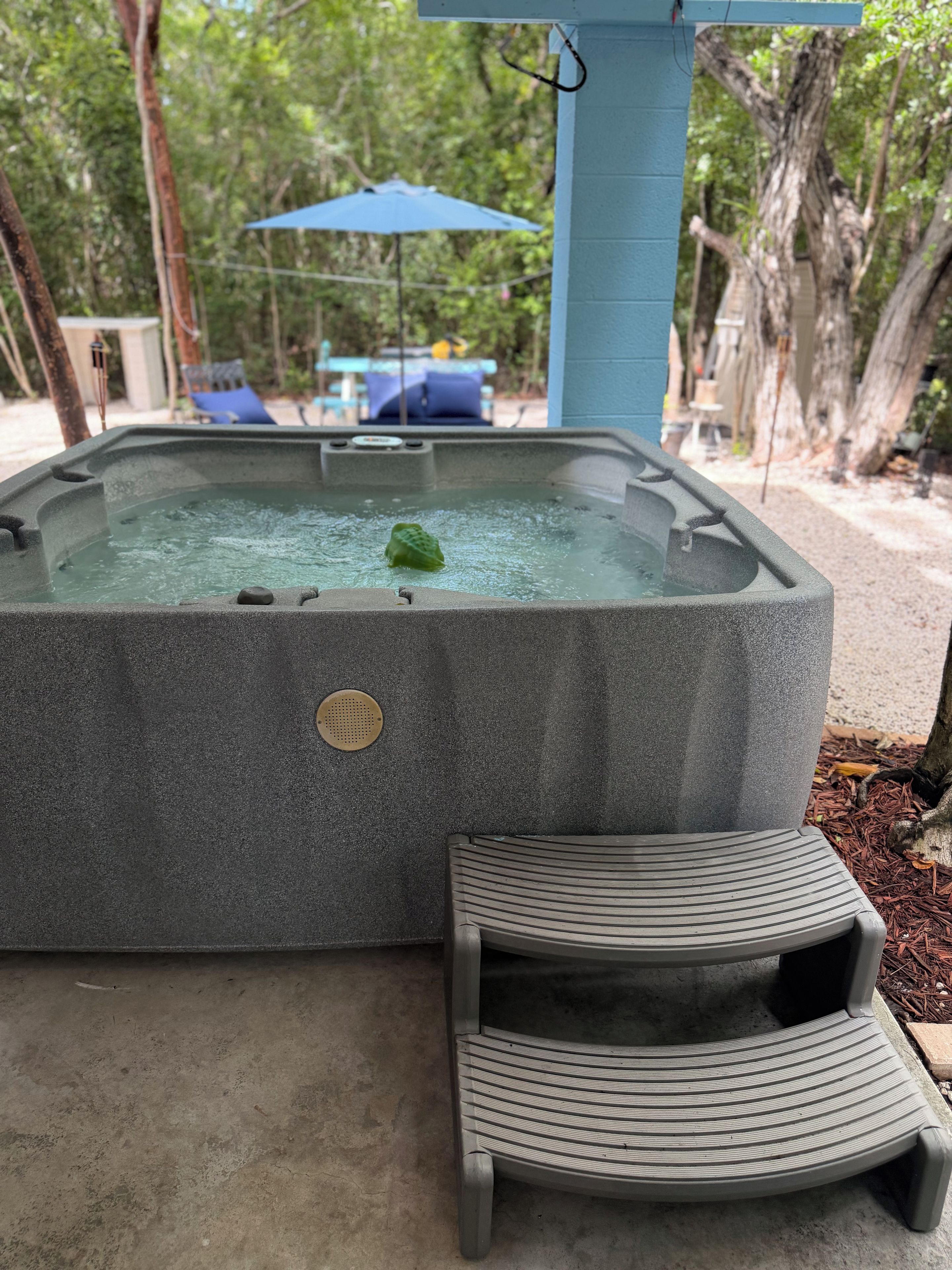 A hot tub with a built-in seat and a small step leading into it, surrounded by a concrete deck and a blue umbrella in the background.