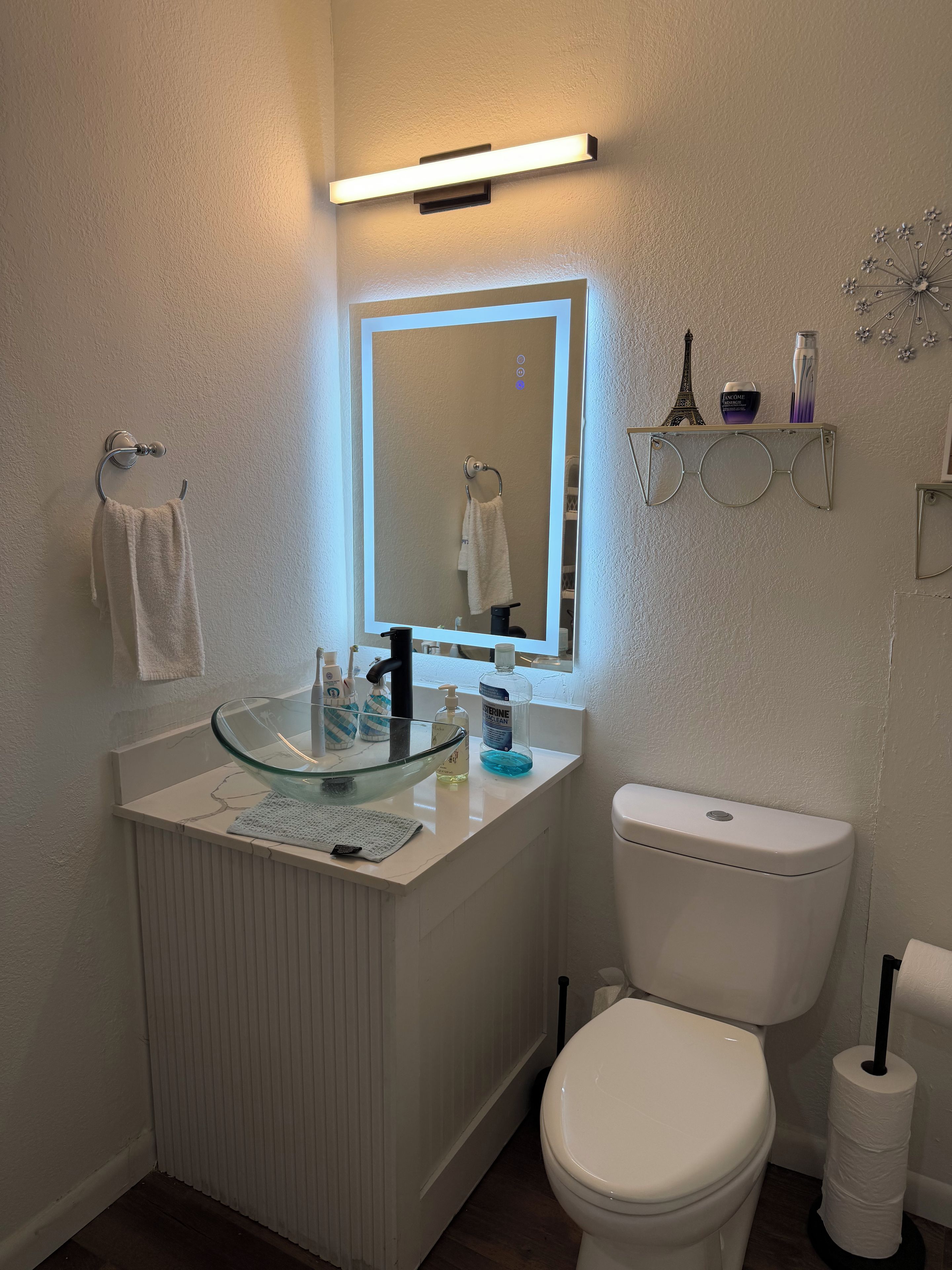 Un baño con un lavabo de vidrio, un inodoro blanco y un espejo con luz superior.